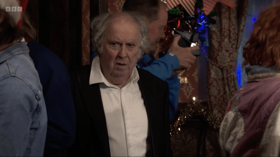 An older man in a black jacket and white shirt looking toward the camera with a confused or displeased expression.