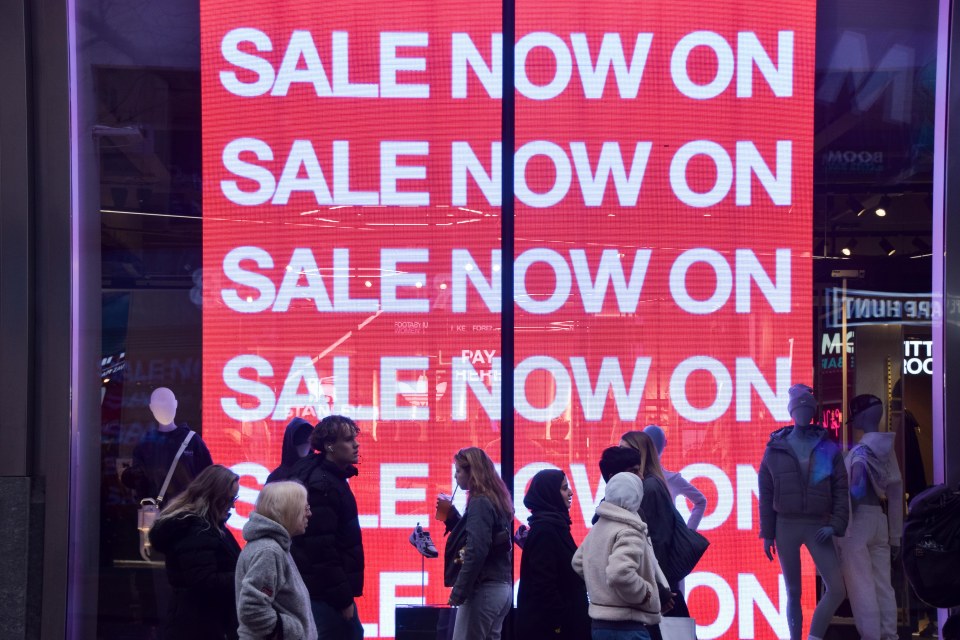 People walk past a store on Oxford Street with a bright red sign displaying "SALE NOW ON" in London, UK.