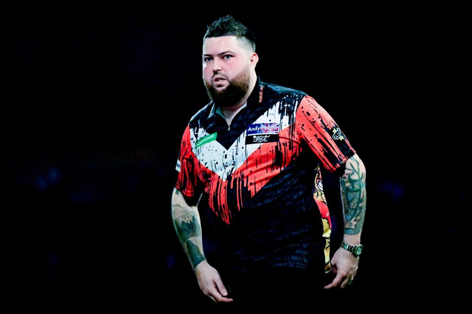 Michael Smith in his darts shirt with sponsor logos, sleeves showing tattoos, at the World Darts Championship.