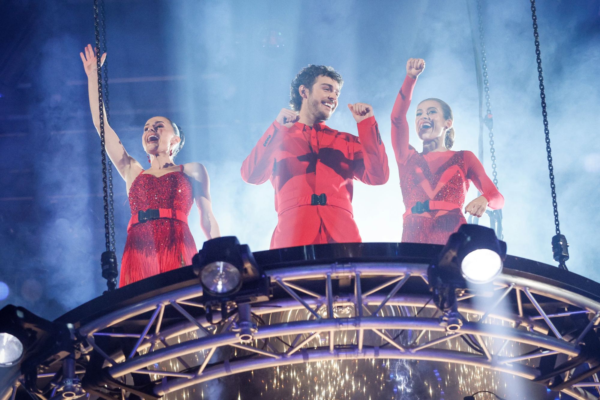 An image collage containing 1 images, Image 1 shows Three dancers, a man and two women, in red costumes celebrate on a raised platform with smoke and sparks