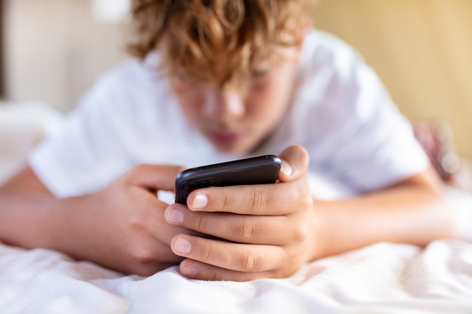 Teenage boy with curly hair lying in bed and looking at a smartphone.