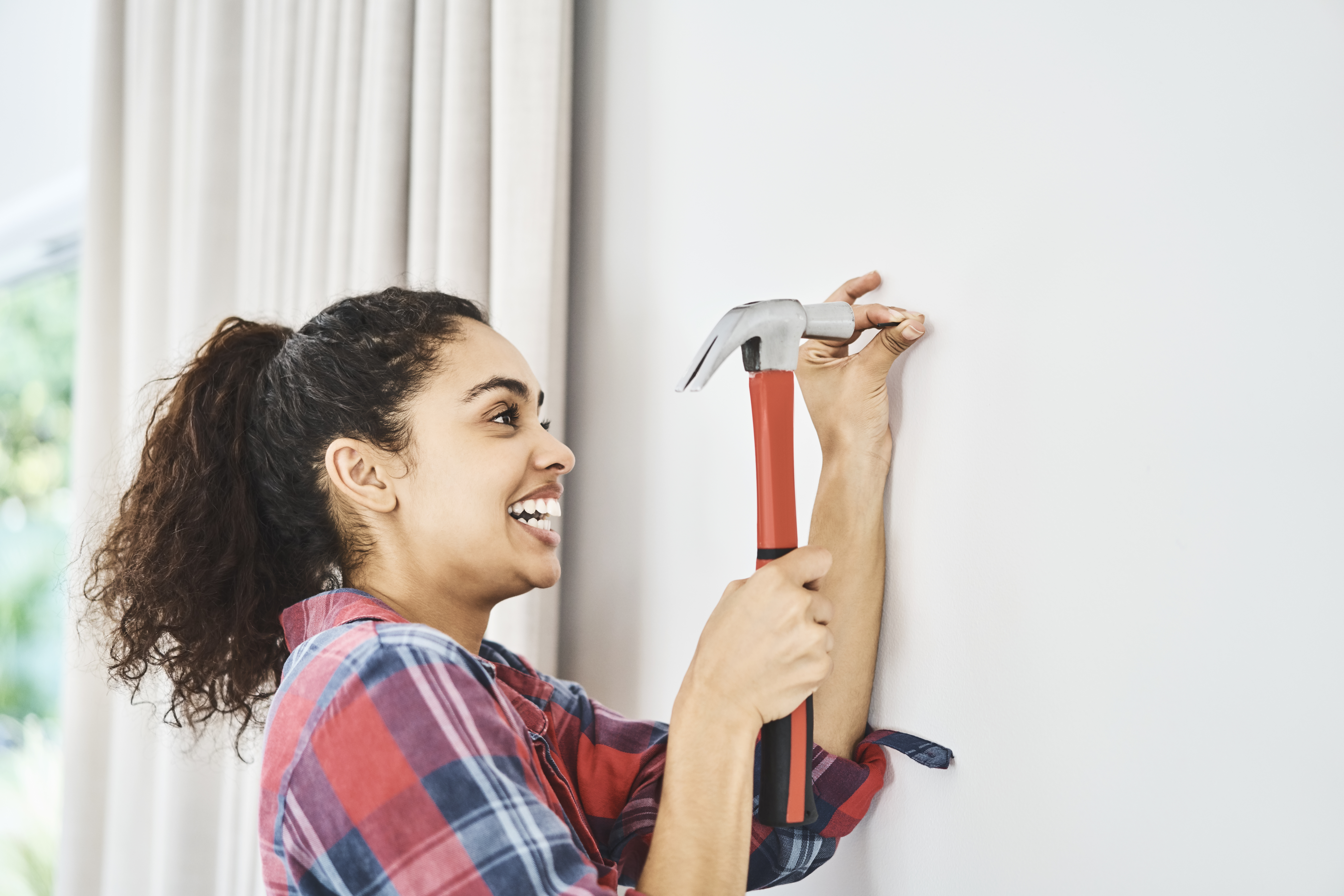 Side view of cheerful young woman hammering nail on white wall. Beautiful female is holding tool in domestic room. She is plaid shirt at home.