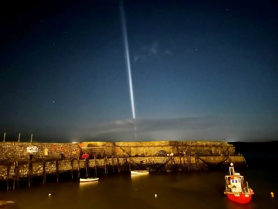 A vertical beam of light extends from a cloudy horizon into the night sky, above a dark harbor with boats and a stone wall.