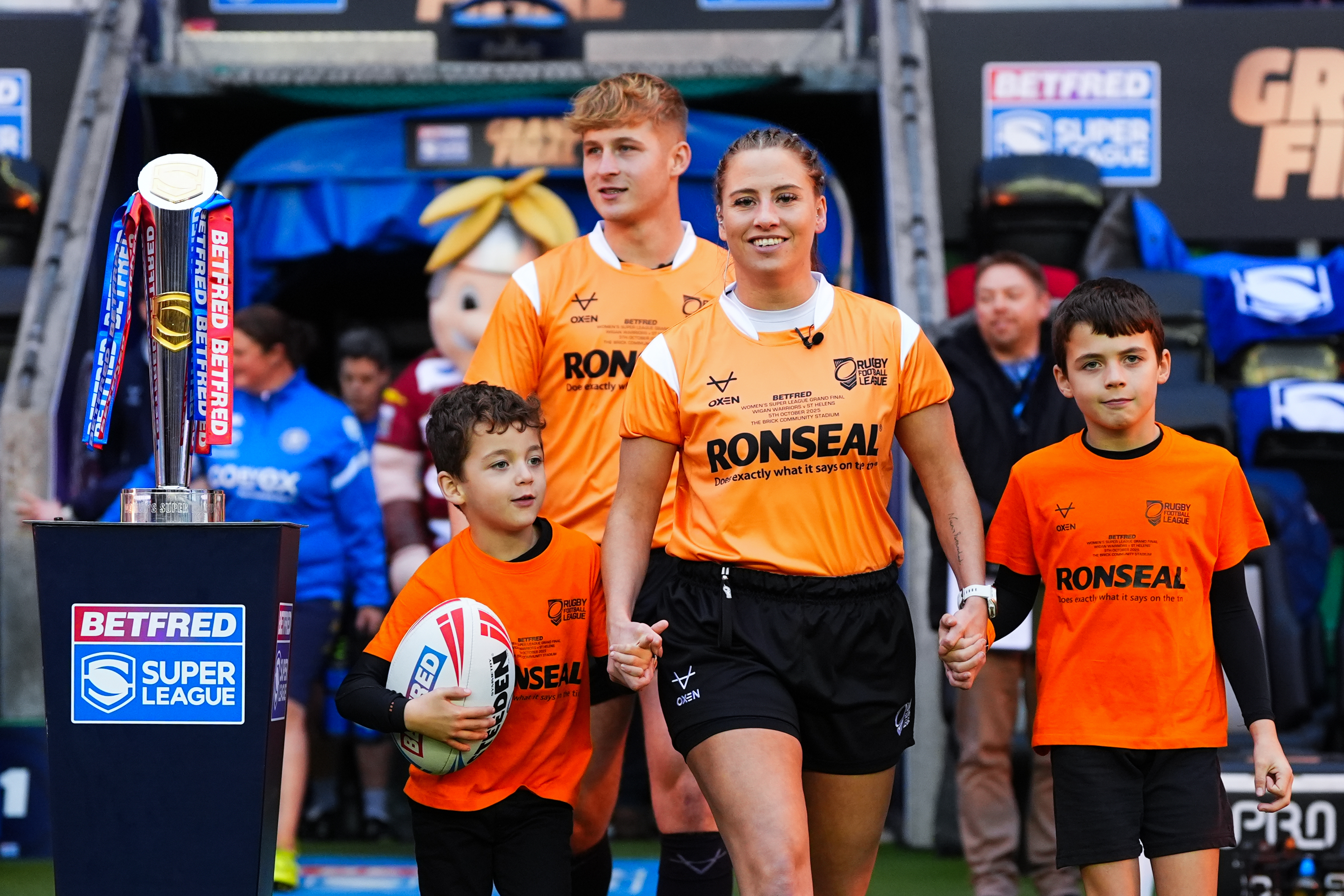 Three officials and two young mascots in orange shirts and black shorts walk out onto a rugby field next to the Betfred Super League trophy.
