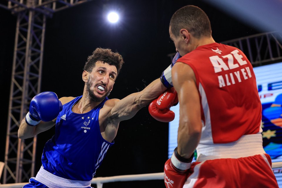 A boxer in a blue uniform with "FFBOXE" and three stars delivering a punch to a boxer in a red uniform with "AZE ALIYEV" on the back.