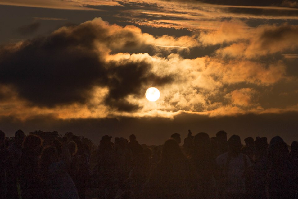 Silhouettes of people in a crowd with a bright setting sun behind clouds.