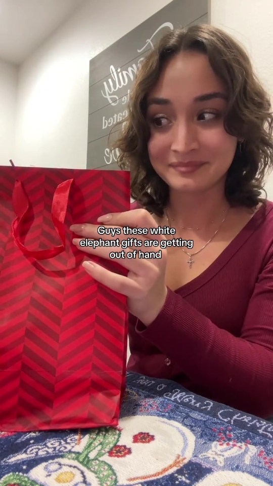 A woman holds a red gift bag with a chevron pattern, looking off-camera with a smirk.