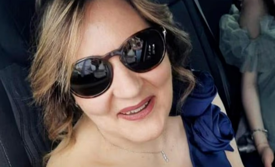 A woman with sunglasses smiling, wearing a blue ruffled top and a silver necklace.