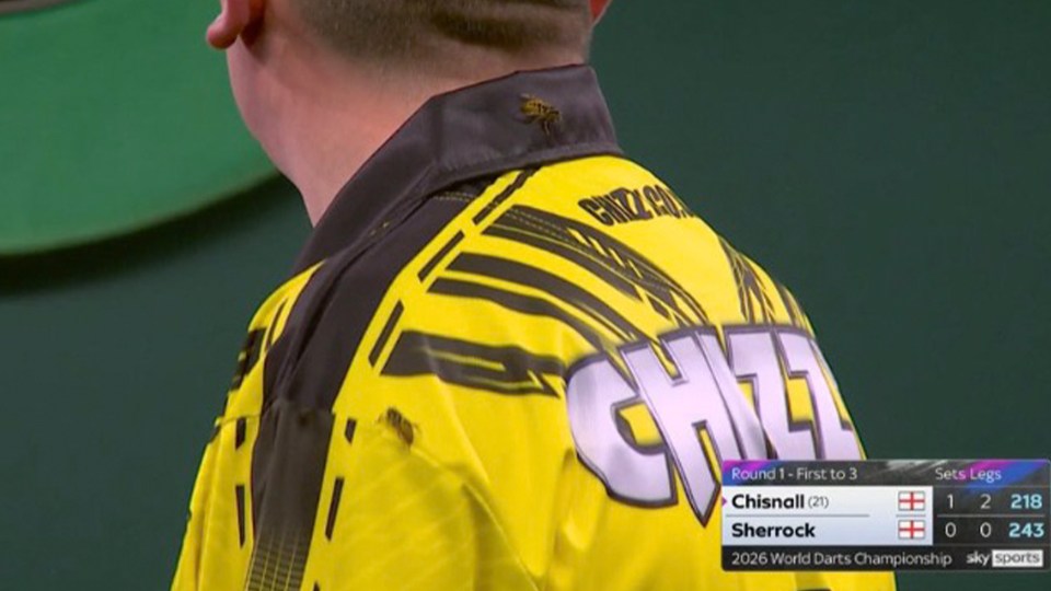 A darts player wearing a yellow and black shirt with a small wasp emblem on the collar, with a scoreboard showing "Chisnall (21)" vs "Sherrock" with scores 1-2 and 0-0 respectively.