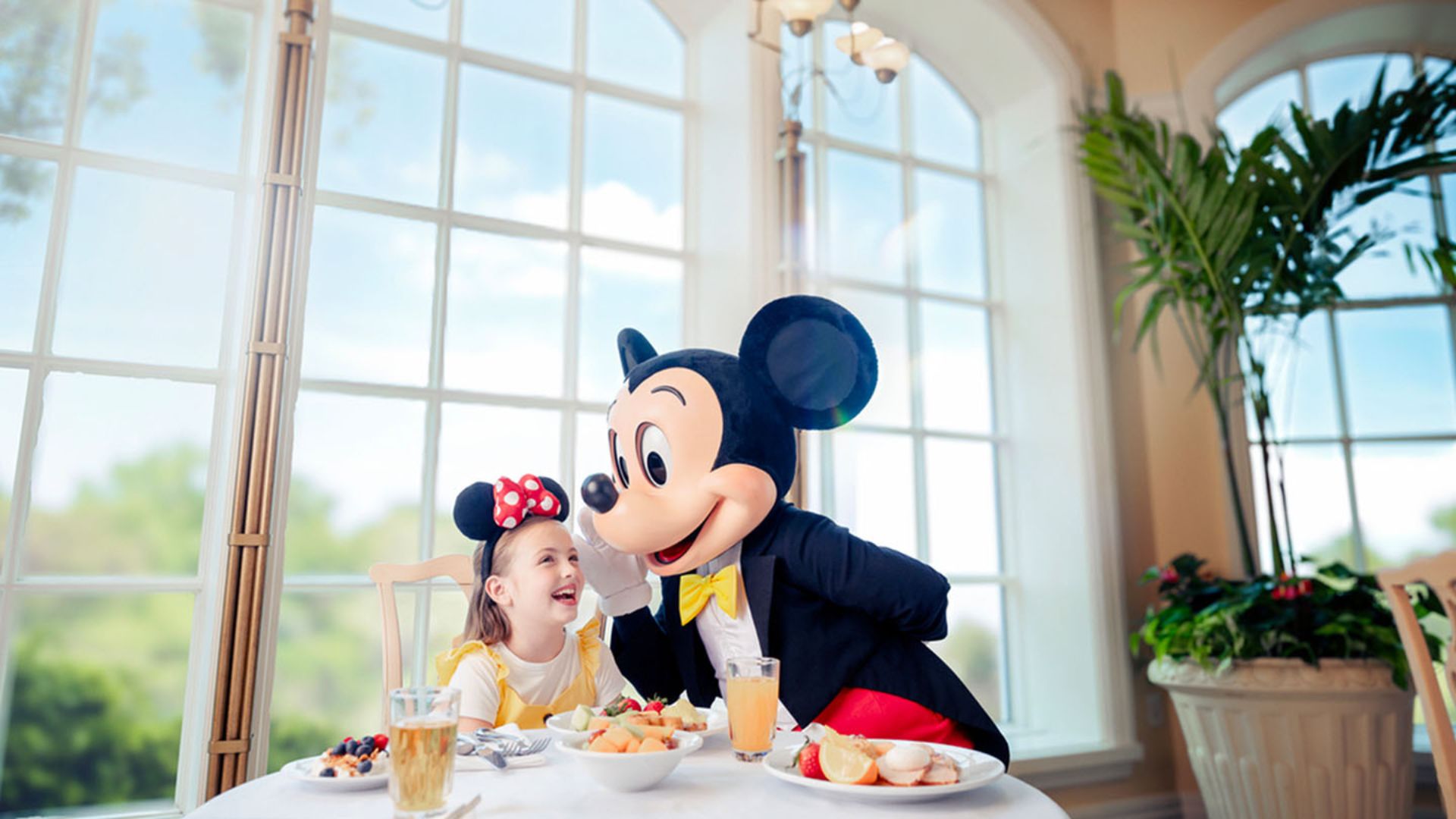 Mickey Mouse mascot sitting with a smiling girl wearing Minnie Mouse ears at a table with breakfast.