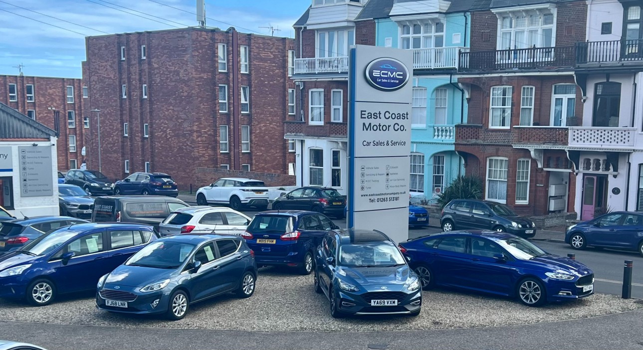 An external view of the East Coast Motor Co. car sales and service center, with various vehicles, a large sign with company information, and buildings in the background.