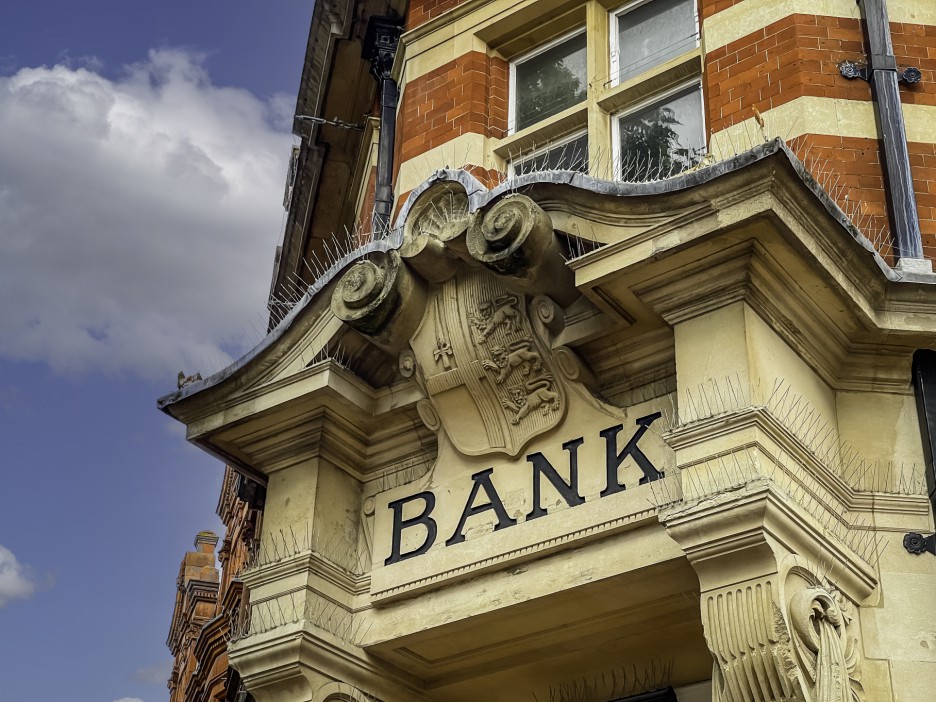 Bank sign with spikes on the ledges, against a brick building and cloudy sky.