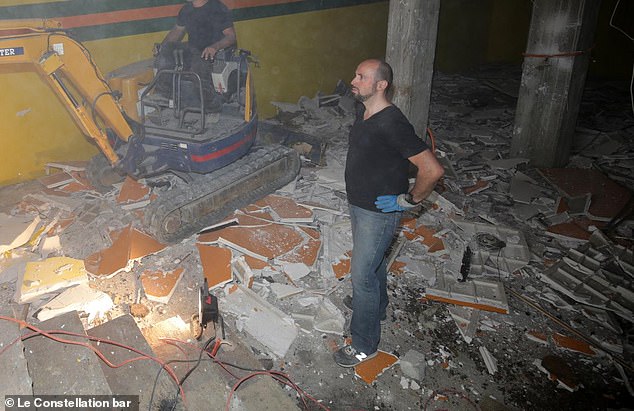 The French owner - who can be seen donning a pair of blue safety gloves - watches on as a man operates a digger amid a sea of rubble the downstairs area of the club which caught fire