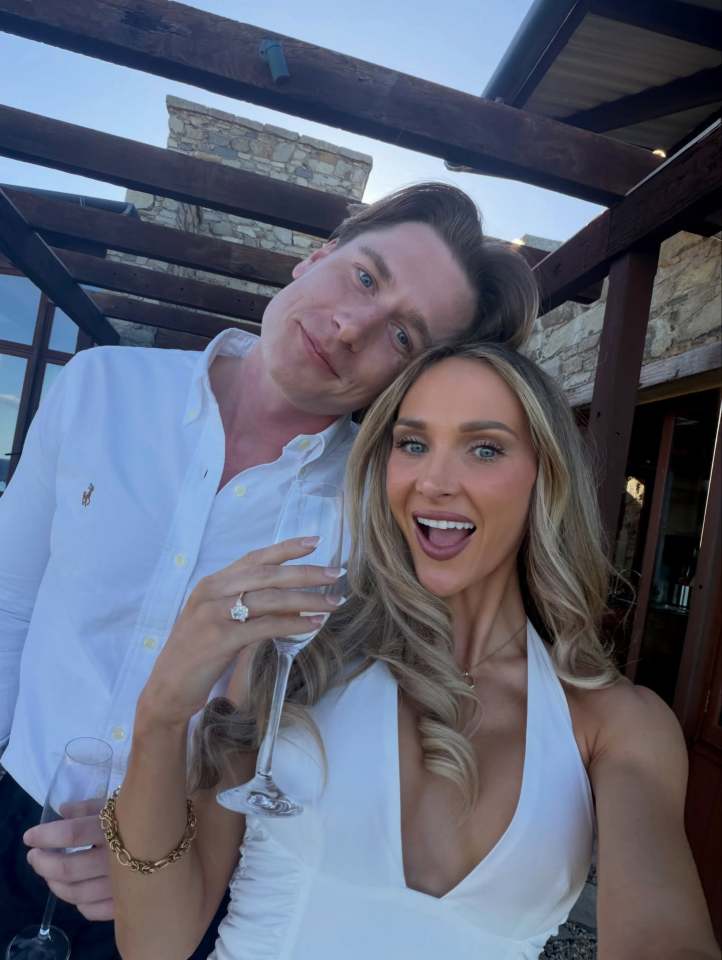 A woman wearing a large engagement ring and holding a champagne glass with a man smiling behind her.