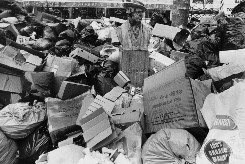 A statue of Reynolds is surrounded by overflowing black bin bags, cardboard boxes, and various waste in Leicester Square, London.