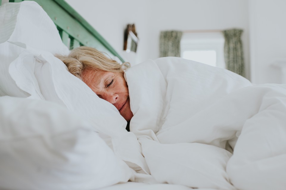 A mature woman snuggles up in a clean, white duvet in a bright and airy bedroom
