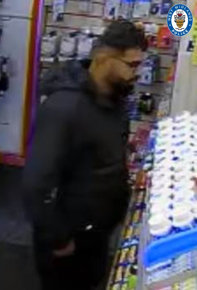 Security footage of a man with dark hair and a beard, wearing glasses and a black hoodie, looking at products in a store.