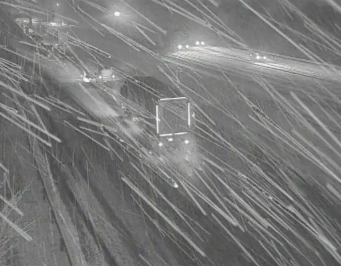 A highway camera view of a snowy road at night, with light trails from vehicle headlights and snow falling across the frame.