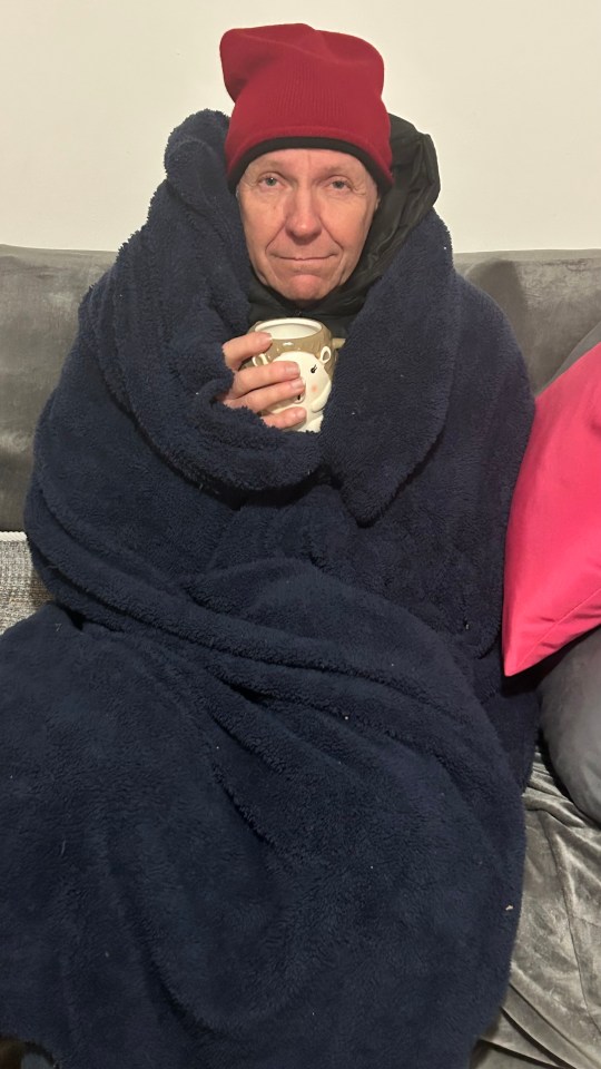 A person sits on a couch, wrapped in a large navy blue blanket with a red hat on their head, holding a mug.