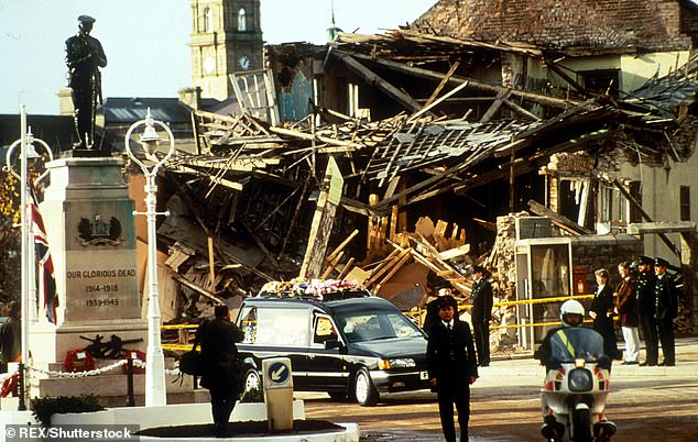 A funeral hearse and police motorcycle drive past the wreckage