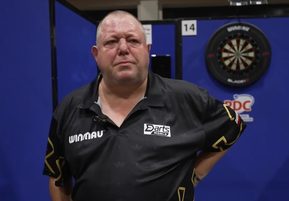 Mervyn King wearing a black Darts World shirt in front of a dartboard.