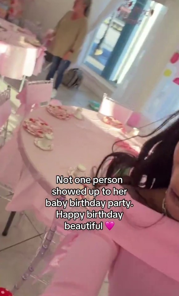 A woman stands in a room decorated for a birthday party with pink tablecloths and chairs, but no guests.