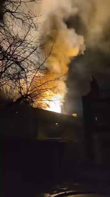 Building engulfed in flames with large plumes of smoke rising into the night sky, framed by bare tree branches.