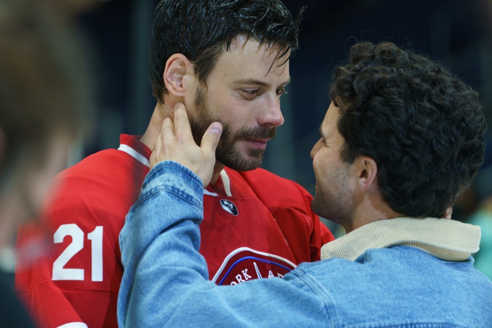 Man in red hockey jersey with number 21 looking at another man in denim jacket who is touching his face.