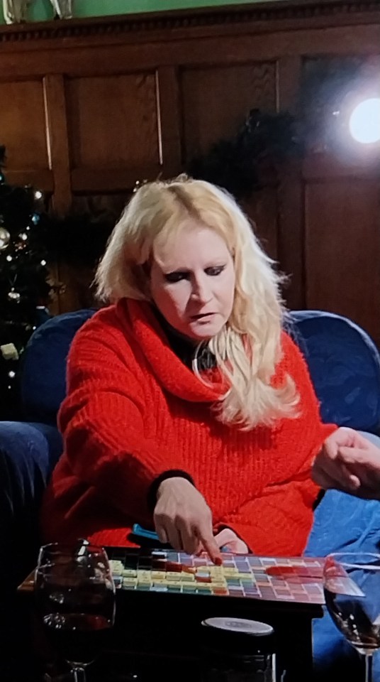 A woman with blonde hair and a red sweater pointing at a Scrabble board.