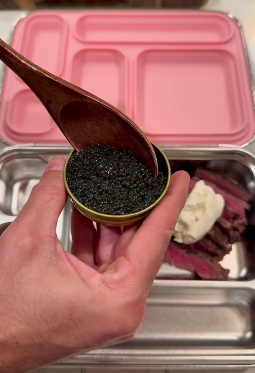 A hand scooping black caviar from a small golden tin with a wooden spoon over a lunchbox containing sliced steak and a dollop of white cream.