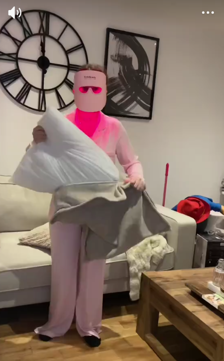 A person in pink pajamas and a pink facial mask putting a pillow into a pillowcase.