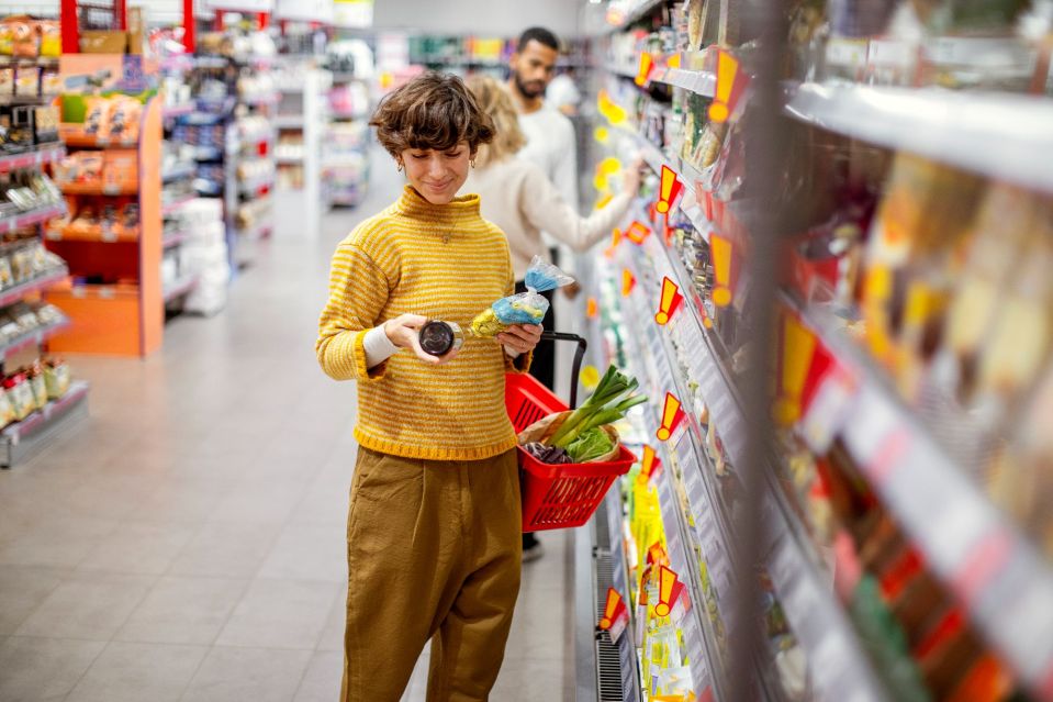 An image collage containing 1 images, Image 1 shows Young woman in a yellow turtleneck examines product labels in a supermarket aisle, holding a red shopping basket with groceries