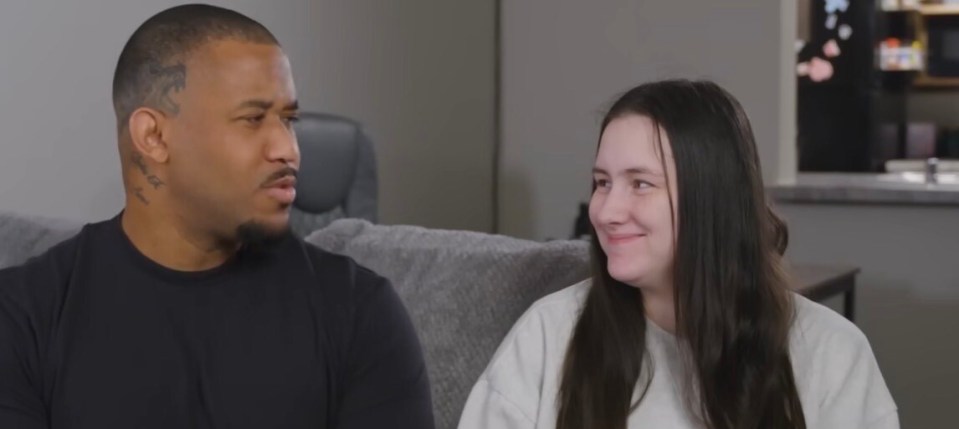 A man with a serious expression and a smiling woman sitting on a couch.