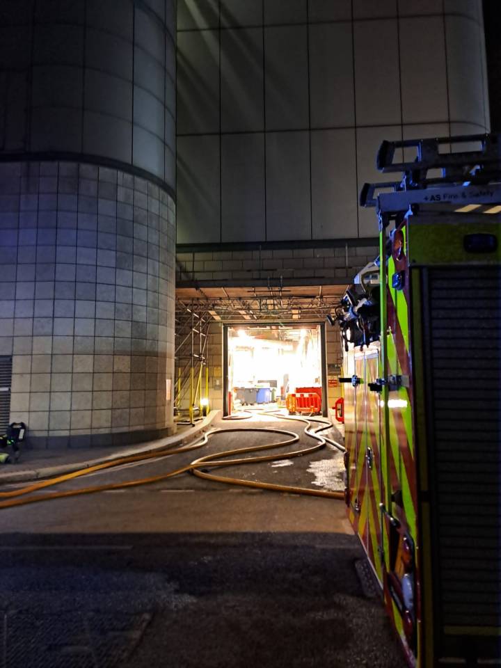 Fire hoses lead from a fire engine towards a brightly lit open doorway in a London tower block, where a fire is being tackled.