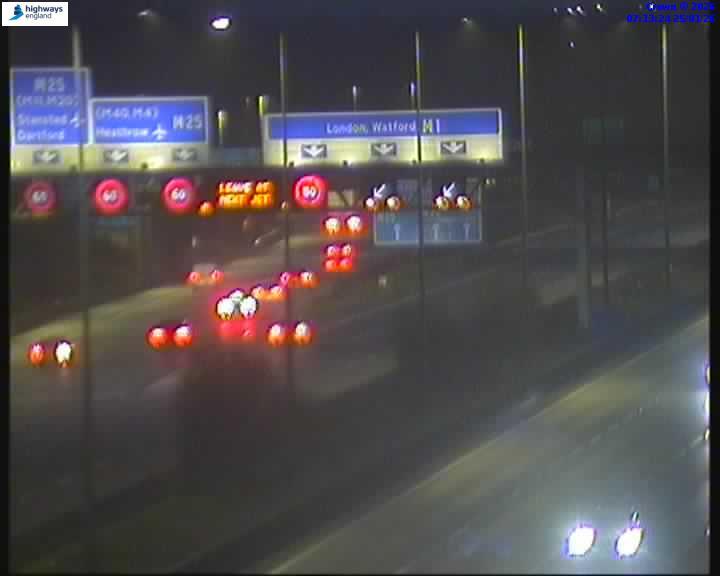 Overhead view of a highway at night, showing traffic with illuminated red taillights and white headlights, along with overhead signs indicating directions and speed limits.