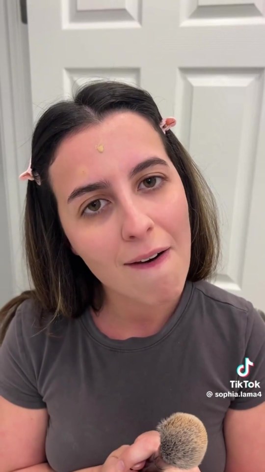 A woman with dark hair, foundation on her forehead, and a makeup brush in her hand, talking to the camera.