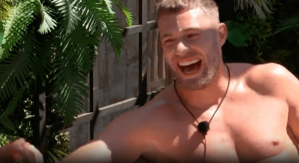 A man with short hair and a beard, wearing a microphone and no shirt, laughs with his mouth open.