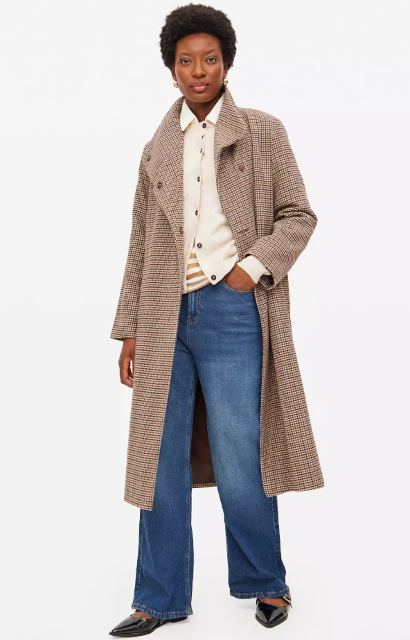 A woman stands wearing a plaid funnel-neck coat, a light-colored button-up shirt, a cream cardigan, wide-leg blue jeans, and black pointed-toe flats.