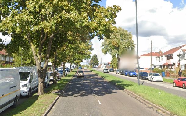 A suburban street with houses on the right, cars parked on both sides, and trees lining the road.