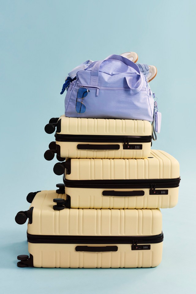 Three light yellow suitcases stacked, with a light purple duffel bag, sneakers, and sunglasses on top.