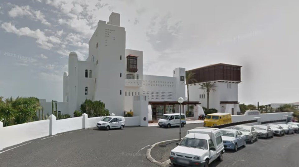 White multi-story building with a brown wooden balcony, surrounded by cars on a street.