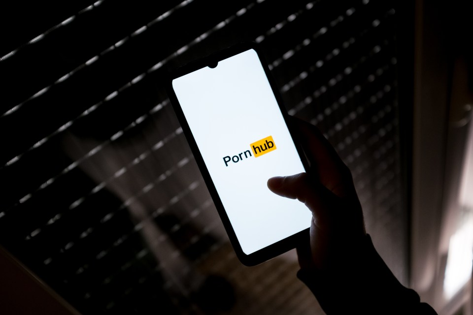 A smartphone displaying the Pornhub logo is held in a silhouetted hand against a dark, striped background.