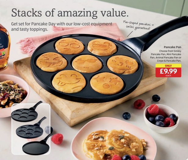 A pancake pan with seven smiley face pancakes cooking in it, next to a bowl of nuts and seeds, and a plate of cooked pancakes with berries.