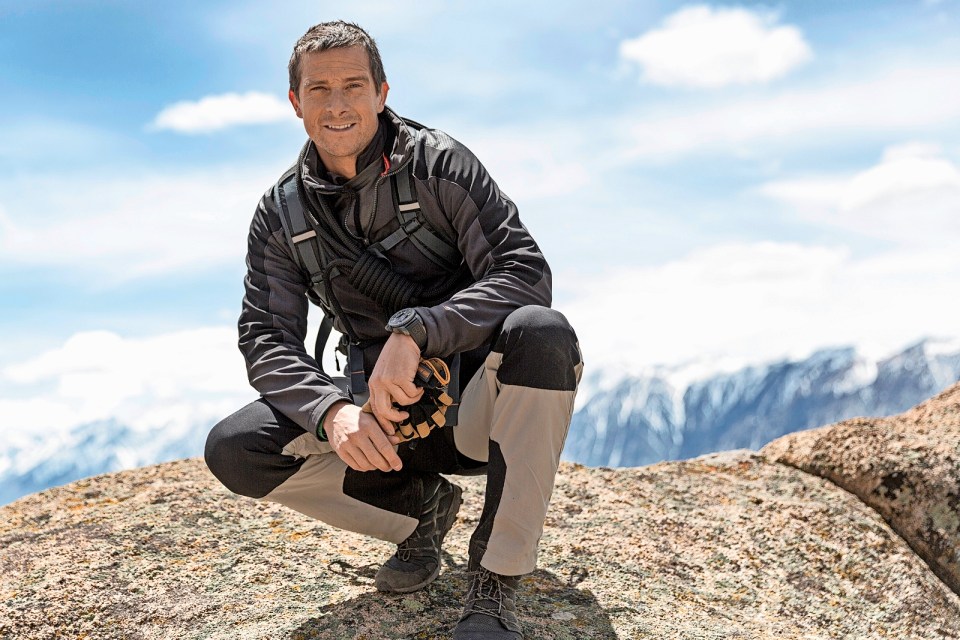 Bear Grylls crouches on a rocky outcrop in front of mountains.