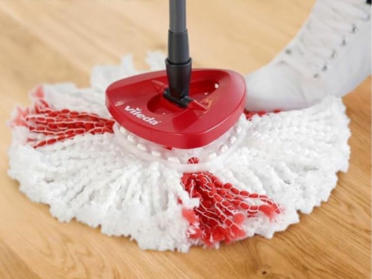 A Vileda spin mop with red and white strands on a wooden floor, with a white shoe visible in the background.