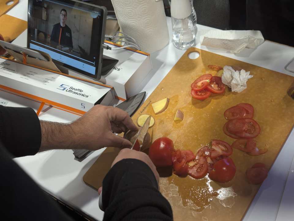 A person cutting vegetables on a brown cutting board, with a tablet playing a video visible in the background.