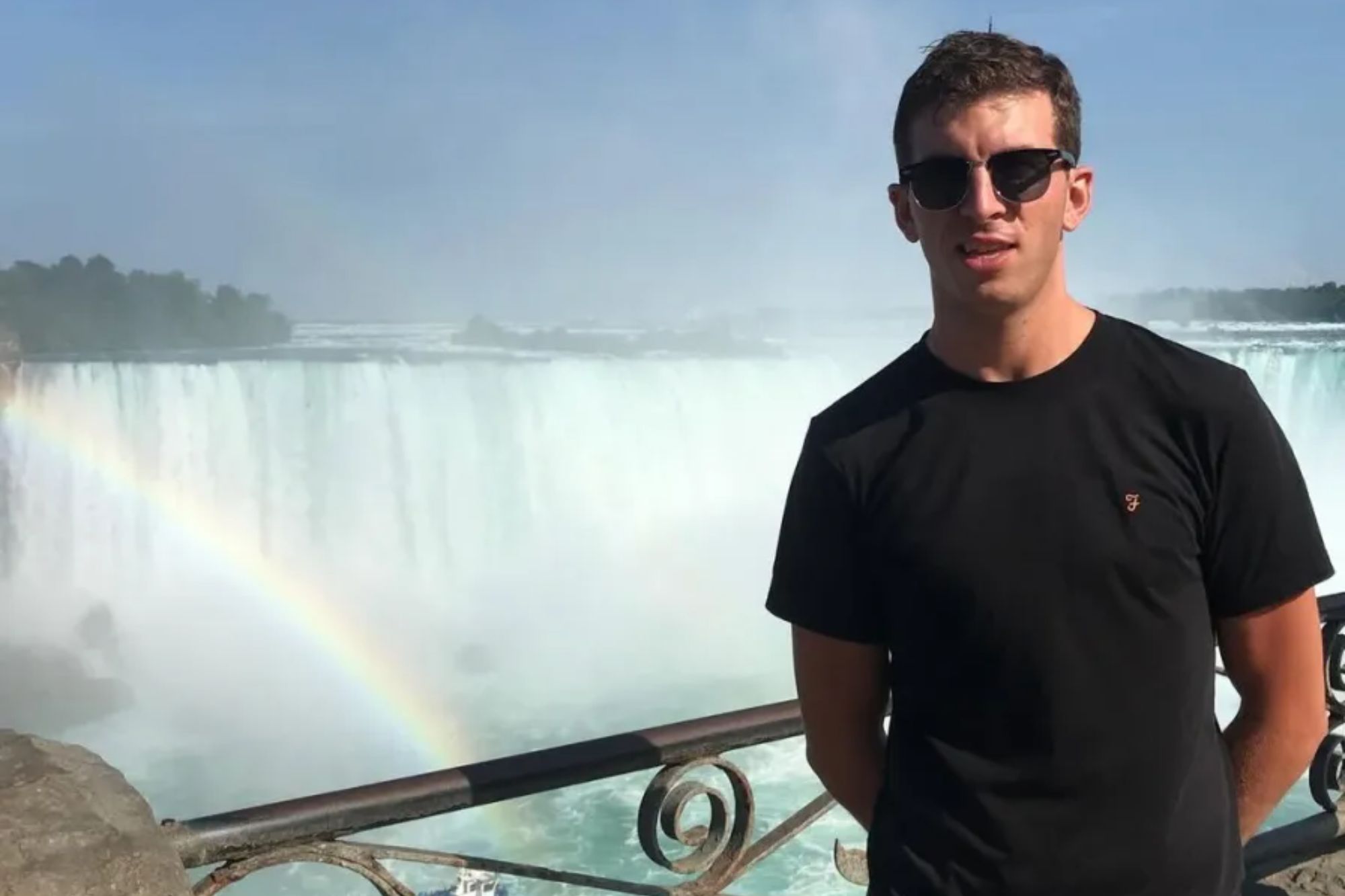 An image collage containing 1 images, Image 1 shows Man in sunglasses and black shirt standing in front of Niagara Falls with a rainbow and a boat below