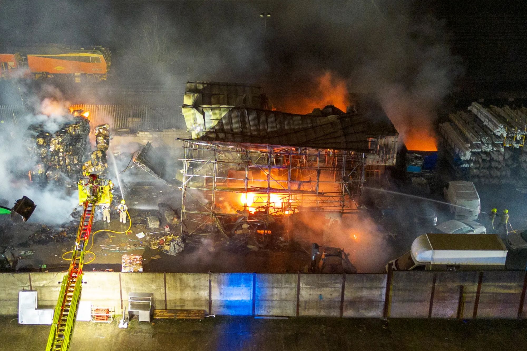 An image collage containing 1 images, Image 1 shows Firefighters battling a large fire at a recycling center with huge plumes of smoke and visible flames