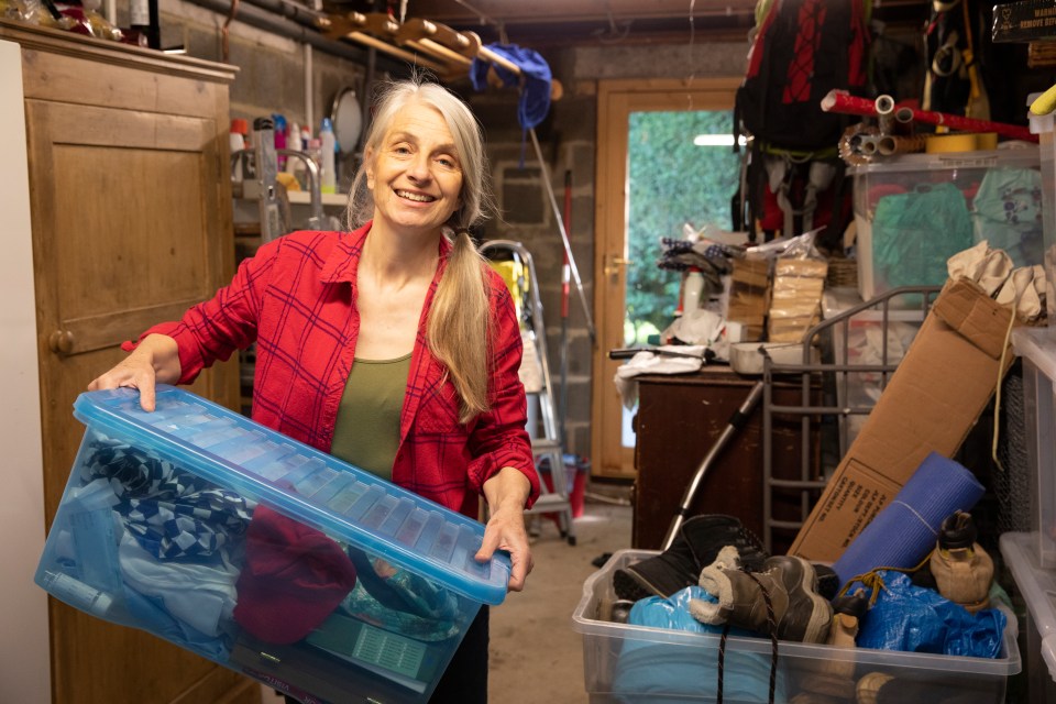 A mature woman smiles while carrying a clear plastic box of belongings in a storage room.