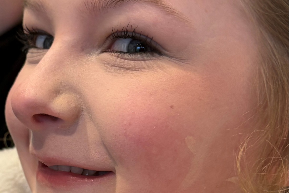 A young girl with blonde hair smiling while wearing makeup, including eyeliner and blush.